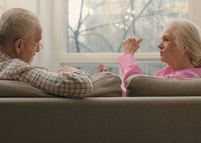 A mature man and woman sit on a tan couch in a living room discussing retirement spending.