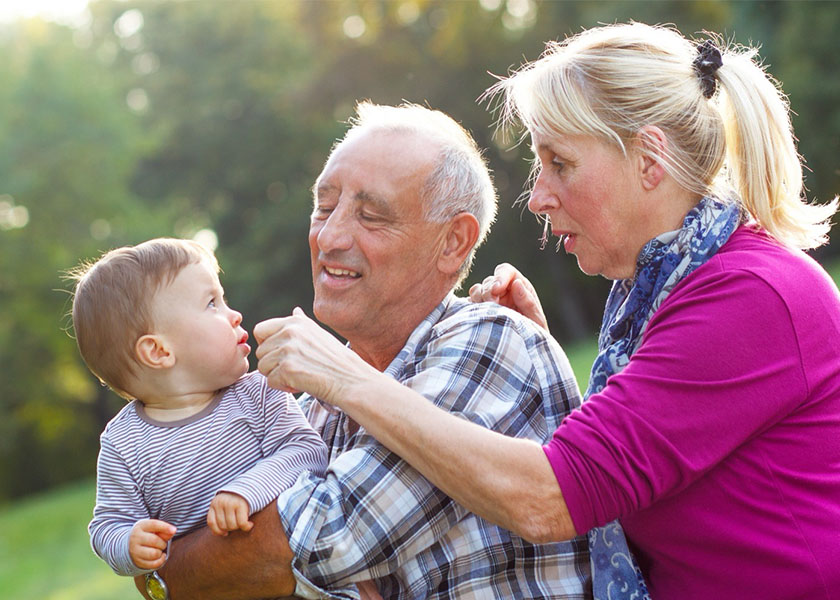 A mature man standing outdoors holding his toddler grandson, smiling, while his wife interacts with them. Finding your financial freedom.