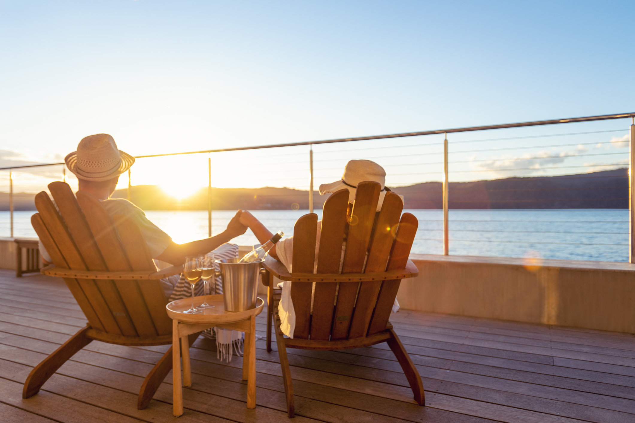 Couple relaxing and drinking wine and holding hands on deck chairs in an over water bungalow. They are looking at the view.